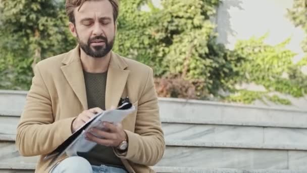幸せなエレガントなひげを生やした男でコートを読んでドキュメントと何かを書きながら 階段の屋外に座って — ストック動画