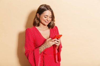 Kırmızı elbiseli, gülümseyen ve cep telefonunu bej arka planda tutan genç bir kadının fotoğrafı.