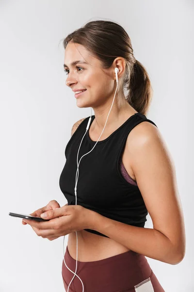 スマートフォンを持ち 白い背景にイヤホンで音楽を聴くスポーツウェアの若い女性のイメージ — ストック写真