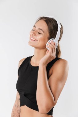 Spor giyimli, gülümseyen ve kulaklıklı, beyaz arka planda izole edilmiş müzik dinleyen atletik bir kadın.