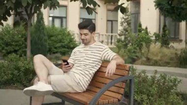 Beyaz bir adam gülümsüyor ve cep telefonunu şehir caddesinde bankta otururken kullanıyor.