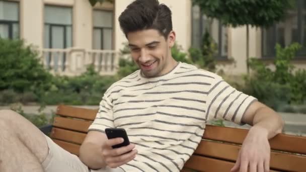 Homem Bonito Está Sorrindo Usando Celular Enquanto Está Sentado Banco — Vídeo de Stock