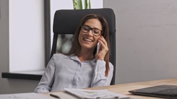 スマートフォンで話す眼鏡をかけ オフィスの椅子でリラックスした幸せなビジネス女性 — ストック動画