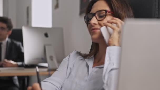 オフィスに座っている間にスマートフォンで話している眼鏡をかけているビジネスマンの女性 — ストック動画