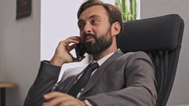 Resmi takım elbiseli mutlu iş adamı akıllı telefonla konuşuyor ve masada otururken yüzünü çeviriyor.