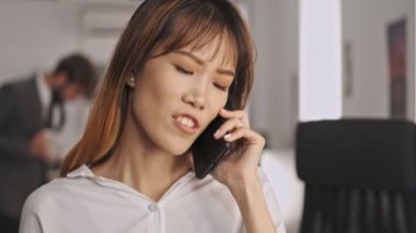 Gülümseyen Asyalı iş kadını akıllı telefonla konuşuyor ve ofiste dikilirken gözlerini kaçırıyor.