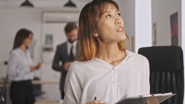 忧心忡忡的亚洲女商人拿着写字板 一边站在办公室一边 一边望着别处 — 图库视频影像