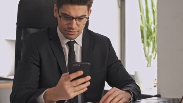 坐在办公室桌子旁 用智能手机和手提电脑近距离观看身着正装 戴着眼镜的严肃商人 — 图库视频影像