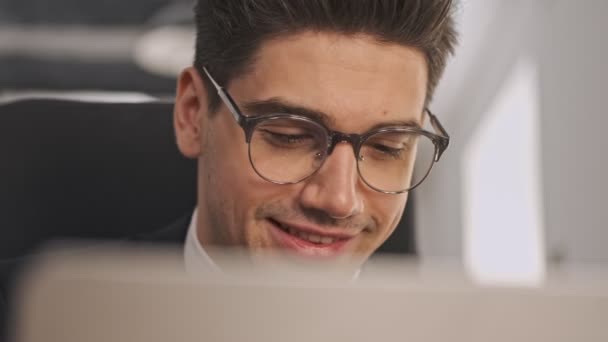 オフィスに座っている間 ラップトップコンピュータを使用して正式なスーツや眼鏡で陽気なビジネスマンのビューを閉じます — ストック動画