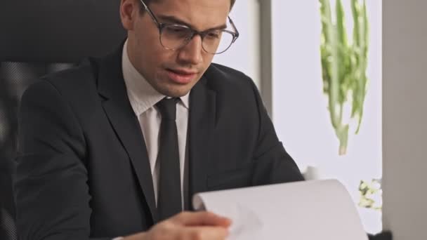 穿着正式西装和眼镜的专心致志的商人坐在办公室的桌子边 一边用剪贴板研究文件 — 图库视频影像