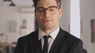 Resmi takım elbiseli ve gözlüklü gülümseyen işadamı ofiste dururken kameraya bakıyor.