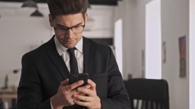 Resmi takım elbiseli ve gözlüklü ciddi bir işadamı ofiste dururken akıllı telefon kullanıyor.