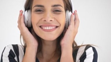 Kulaklık takmış, müzik dinleyen ve gri arka planda kameraya bakan neşeli, güzel amigo kadının yakın görüntüsü.