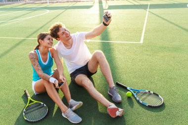 Çekici, sağlıklı tenis oyuncuları stadyumdaki çimlerde otururken cep telefonlarıyla selfie çekiyorlar.