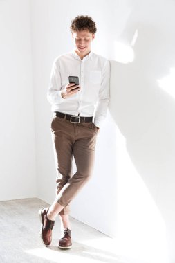 Başarılı, genç, kıvırcık saçlı ve gülümseyen bir iş adamının tam boy portresi beyaz arka planda duruyor, cep telefonu kullanıyor.