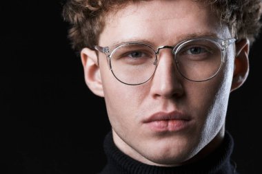 Yakışıklı, kendine güvenen, kıvırcık saçlı, balıkçı yaka kazak giyen, gözlüklü, siyah arka planda poz veren bir işadamının yakın çekimi.