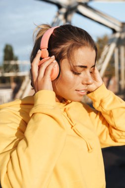 Gülümseyen genç bir fitness kadınının dışarıda poz verip kulaklıkla müzik dinlemesi..
