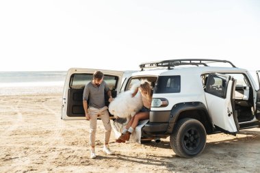 Kumsaldaki neşeli genç çift, köpekleriyle oynuyorlar, arabanın içinde oturuyorlar.