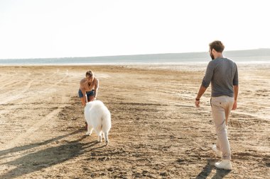 Kumsalda yürüyen neşeli genç çift köpekleriyle oynuyorlar.
