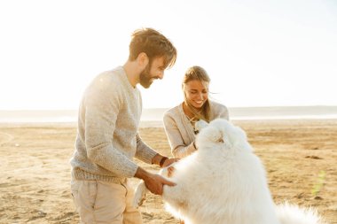 Kumsalda yürüyen neşeli genç çift köpekleriyle oynuyorlar.