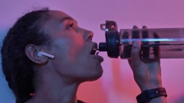 Kulaklıklı, memnun Afrikalı spor kadının duvarın yanında izole edilmiş el fenerleriyle otururken müzik dinlediği ve şişeden su içtiği görüntüyü kapatın.
