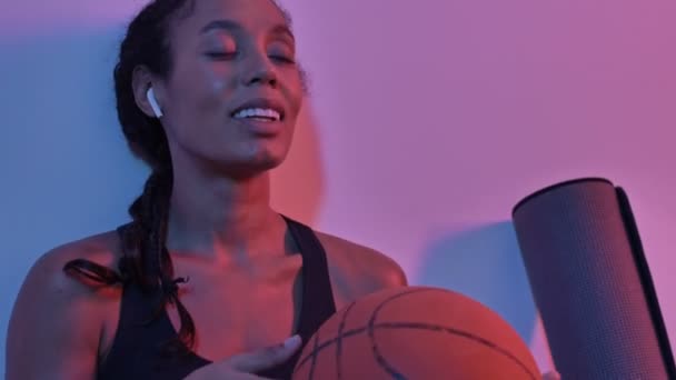 Close up view of pleased african sports woman in earphones listening music and playing with ball while sitting near the wall isolated led flashlights background — Stock Footage Close View Pleased African Sports Woman Earphones Listening Music Playing — Stock Video