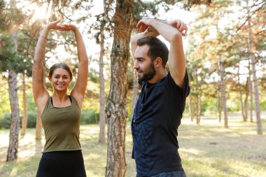 Photo of young cheerful couple in sportswear doing exercises while working out in green park during sunny summer day clipart