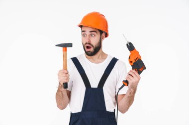Kafasında matkap ve çekiç tutan beyaz duvar arkasında yalnız poz veren şaşırtıcı genç adam görüntüsü.