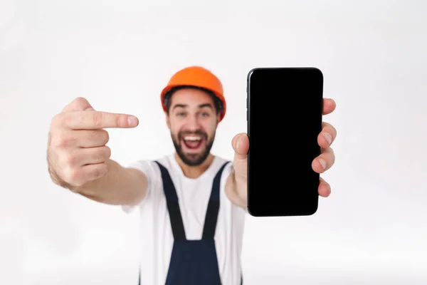 在白色墙壁背景下隔离的头盔中的情绪积极的年轻男性建设者形象 显示了手机指向的显示 — 图库照片