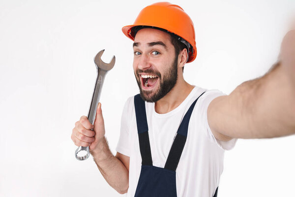 Image of happy positive young man builder in helmet isolated over white wall background take selfie by camera holding wrench.