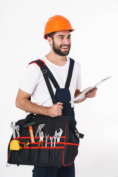 照片中 年轻快乐的建筑工人戴着安全帽 带着装备工具 带着剪贴板 隔离在白墙背风袋上 — 图库照片