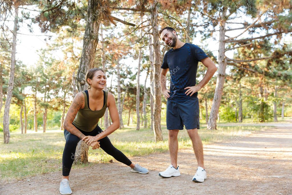 Photo of attractive laughing couple in sportswear doing exercises while working out in green park during sunny summer day