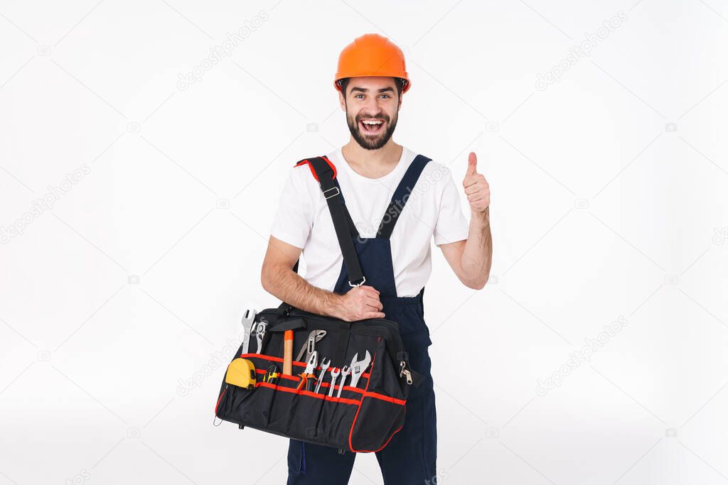 Image of happy smiling young man builder in helmet isolated over white wall background holding bag with equipment instruments showing thumbs up.