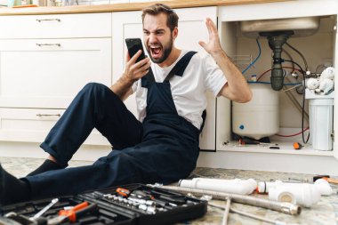 Hoşnutsuz genç bir tesisatçı resmi. İçeride üniformayla çalışıyor. Cep telefonuyla konuşuyor..