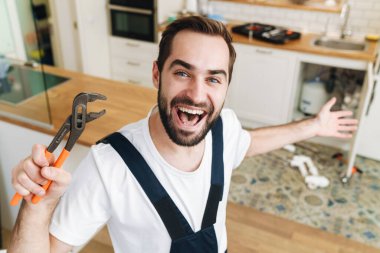 Boru anahtarı tutan, üniformalı, neşeli, genç bir tesisatçı resmi.