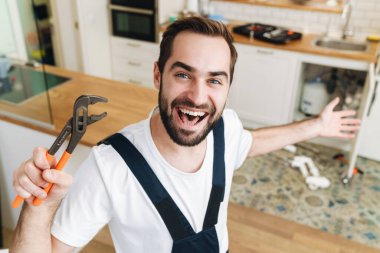 Boru anahtarı tutan, üniformalı, neşeli, genç bir tesisatçı resmi.