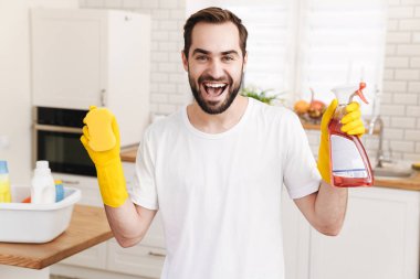Evde elinde sünger ve temizlik spreyi tutan genç, pozitif bir erkek resmi..