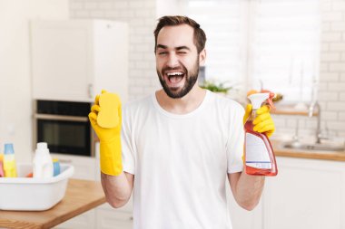 Neşeli, gülümseyen, pozitif bir adamın ev erkeği elinde sünger ve temizlik spreyi tutarken görüntüsü..
