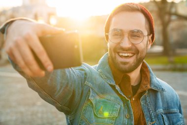 Yakışıklı, mutlu, genç, şık sakallı bir adamın sokakta yürürken selfie çekmesini.