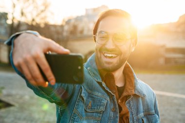 Yakışıklı, mutlu, genç, şık sakallı bir adamın sokakta yürürken selfie çekmesini.