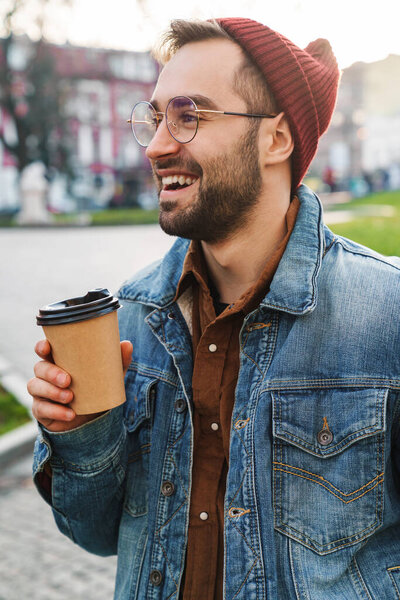 Close up of a handsome happy young stylish bearded man walking outdoors in the street, holding takeaway coffee cup