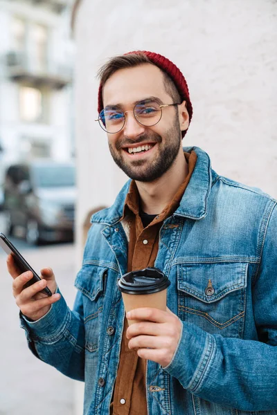 路上を歩くハンサムな幸せな若いスタイリッシュなひげを生やした男のクローズアップ テイクアウトコーヒーカップを保持 携帯電話を使用してください — ストック写真