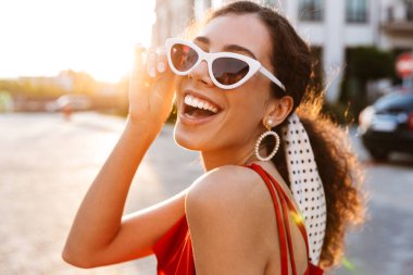 Güneş gözlüklü, güzel, neşeli bir kadının, şehir caddesinde yürürken kameraya bakıp gülüşünün görüntüsü.