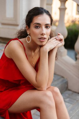 Dışarıdaki merdivenlerde oturmuş kameraya bakan kırmızı elbiseli güzel bir kadın resmi.