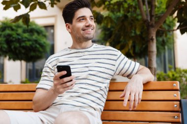 Şehir caddesinde bankta otururken gülümseyen ve cep telefonu kullanan mutlu yakışıklı bir adam.