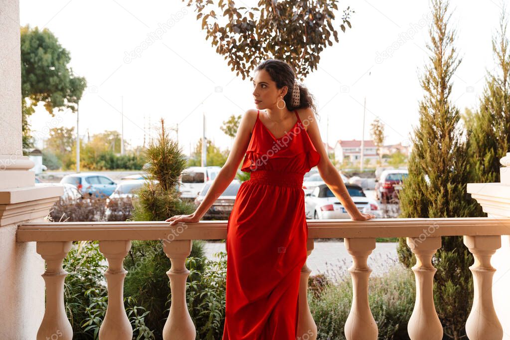 Image of beautiful young woman in red dress looking aside while leaning on railing near colonnades