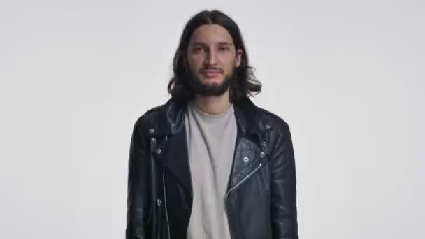 一个穿着黑色皮夹克 留着一头长发的迷人的微笑的年轻人正在白底上眨眼 — 图库视频影像
