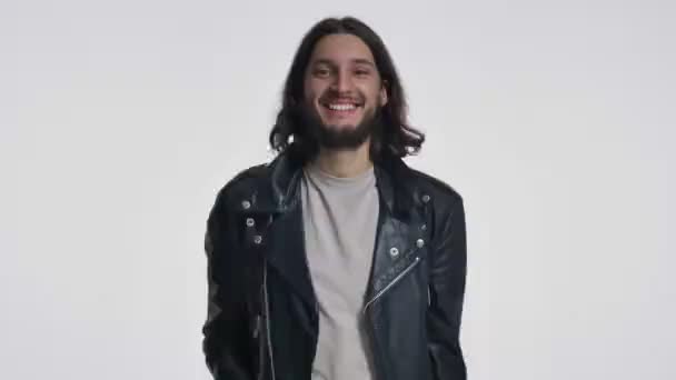 一个穿着黑色皮夹克 留着长发的快乐的年轻人正在向被白色背景隔离的摄像机挥手 — 图库视频影像