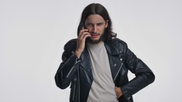 一位穿着黑色皮夹克 留着长发的英俊年轻人在电话里通过白色背景进行着交谈 — 图库视频影像