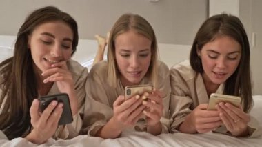 Mutlu genç kız arkadaşlar ışık odasındaki yatakta yatarken akıllı telefon kullanıyorlar.